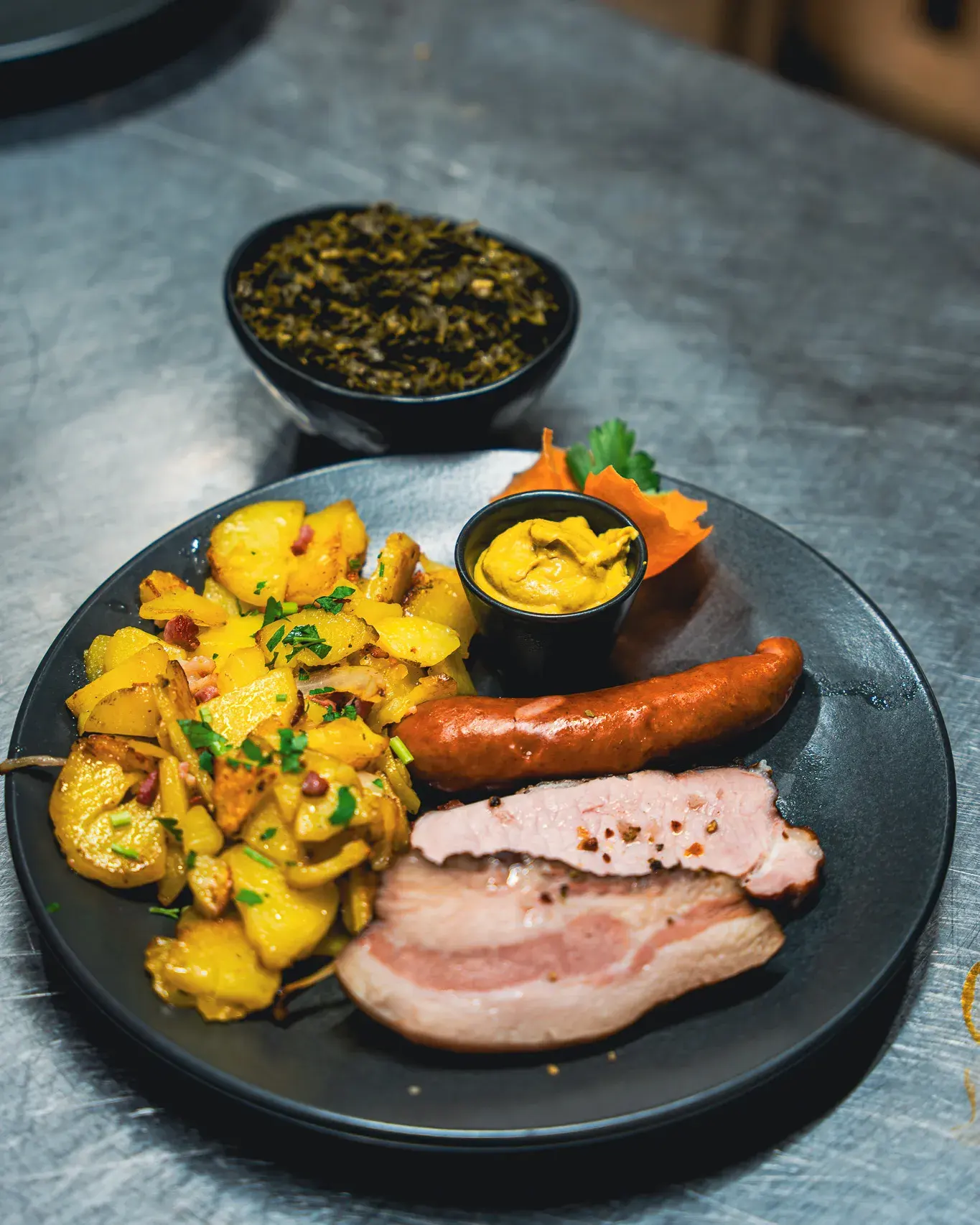 Grünkohl in der Schale, davor ein Teller mit Bratkartoffeln, Kohlwurst, Kassler und Senf