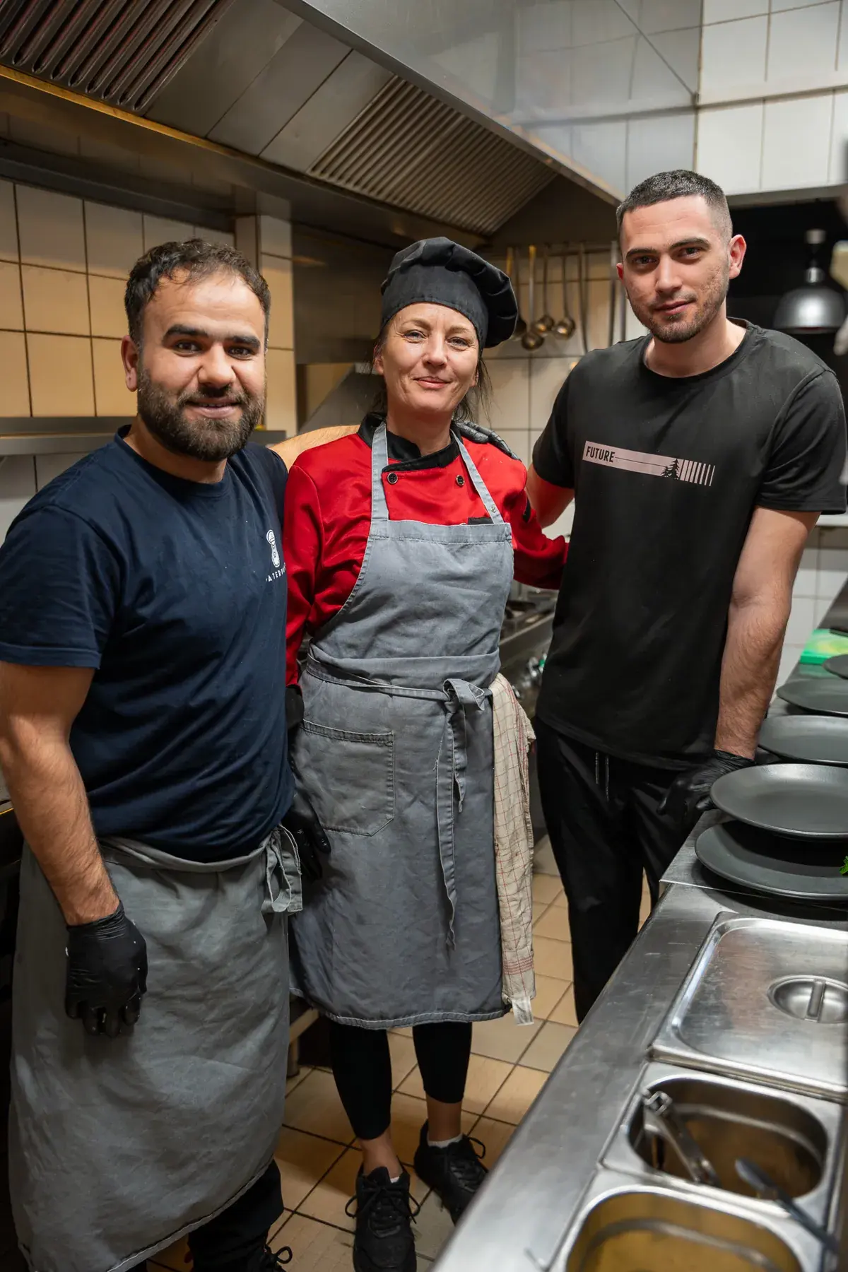 Drei aus dem Küchenteam stehen in der Küche, die Köchin mit Kochmütze in der Mitte