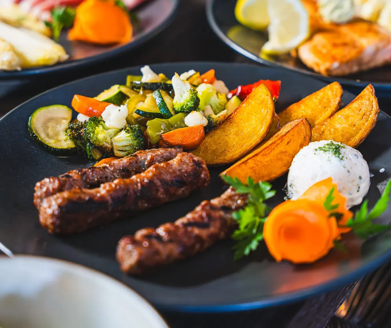 Zwei Cevapcici mit Kartoffelspalten, gebratenem Gemüse und Karottenblüte auf dunklem Teller